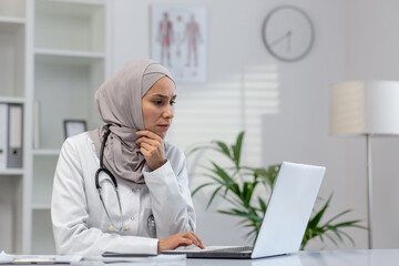 Focused Muslim female doctor, wearing a hijab, sits over her laptop in a modern clinic office, appearing thoughtful and concerned.