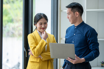 Professional Asian male and female colleagues discuss work on a laptop in a bright modern office, looking engaged and focused.