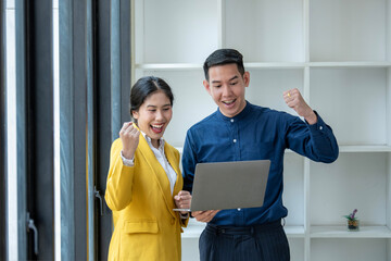 Two Asian business professionals excitedly celebrate success, looking at a laptop screen in a...