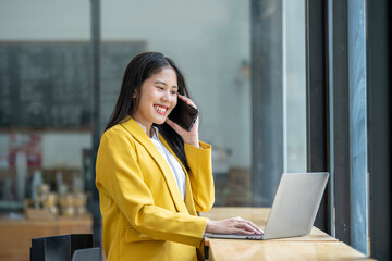 Smiling Asian young woman enjoys using her smartphone while sitting at a modern work desk with a...