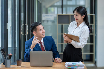 Professional Asian male and female colleagues discuss work on a laptop in a bright modern office, looking engaged and focused.