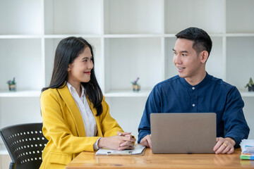 Professional Asian male and female colleagues discuss work on a laptop in a bright modern office,...