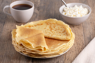 A stack of thin pancakes, a plate with cottage cheese and coffee on a wooden table