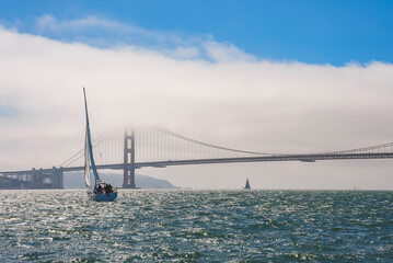 Serene maritime scene with Golden Gate Bridge in San Francisco. Bridge covered in mist, bay waters glisten under sunlight, sailboats voyage leisurely. - Powered by Adobe