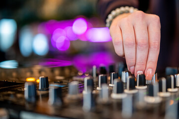 Close-up of a DJ’s hand adjusting knobs on a mixer, adorned with bracelets. Purple lights in the background create a club ambiance - obrazy, fototapety, plakaty