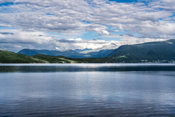 Overlooking the flat, calm waters of the Ålvundfjord. View of the other side of the fjord coastline with the village of Handlarstuhamran against the backdrop of the Trollheimen Mountains in Norway.