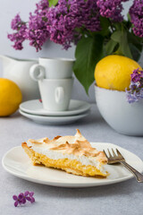 A piece of lemon tart with meringue on a plate, lilac flowers, a whole lemon, coffee cups.