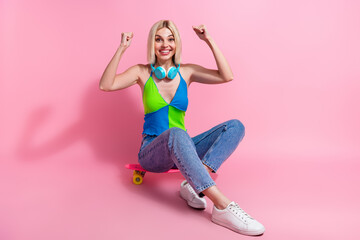 Full size photo of ecstatic woman with headphones sit on skateboard raising fists scream yes win...