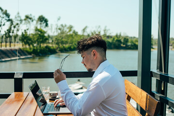 A man with a laptop in a cafe.
A young bearded man in glasses is sitting at a table in a cafe in front of an open laptop by the river - Powered by Adobe