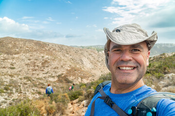 Cheerful male hiker with hat takes a selfie with mountains on background. 