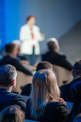 A focused image showing the back of attendees listening to a businesswoman speaking at a corporate...
