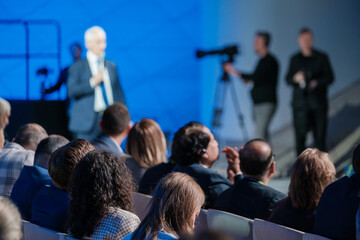 Focused photograph of a senior businessman speaking energetically at a corporate conference. Blurred audience in foreground, capturing attention and interaction.