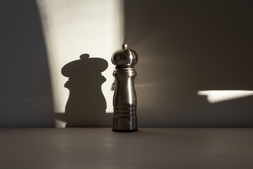 Selective focus of tall stainless steel pepper mill set on table in sun ray with shadow on pale wall