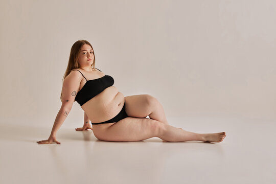 Elegance and confidence. Beautiful young girl with plus size body shape posing on floor in underwear against grey studio background. Concept of natural beauty, body positivity, care, acceptance