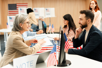 Senior worker woman working at polling station, sitting at registration table, talking to man, voter