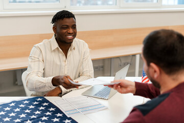 Smiling African American man worker working at polling station, holding ballot, talking to voter