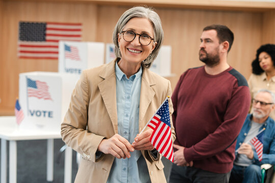 Smiling beautiful senior woman voter wearing eyeglasses holding American flag looking at camera