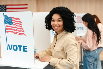 Smiling beautiful African American woman with curly hair standing in booth with American flag