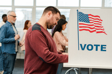 Attractive, middle aged man standing in voting booth with American flag, voting at polling station