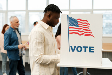 Young African American man standing in voting booth with American flag, voting at polling station