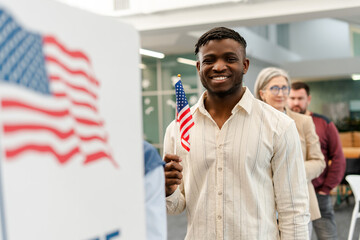Smiling African American young man holding American flag standing at polling station near booth
