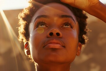 A closeup of a young black woman with a nose piercing, looking focused and pointing upwards, with soft sunlight in the background