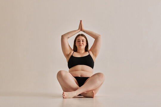 Young woman with plus size body sitting in yoga pose, feeling confident and comfortable in her body against grey studio background. Concept of natural beauty, body positivity, care, acceptance