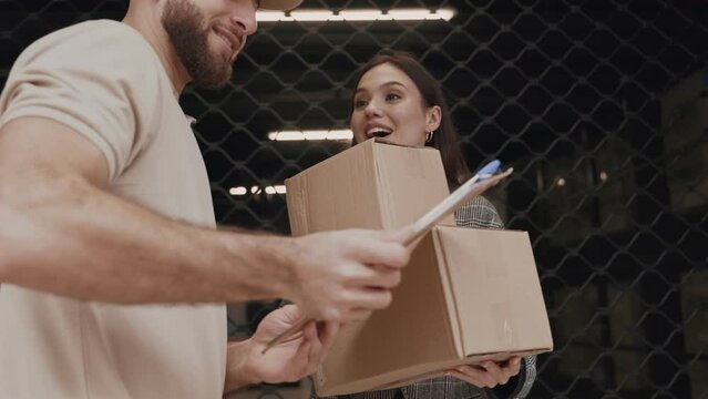 An image showcasing a smiling woman receiving a package from a male courier, who is asking for a confirmation signature. The exchange occurs outdoors, by a mesh fence.