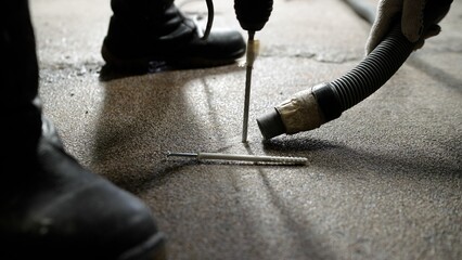 One man drills a hole in the floor with a hammer drill, the second holds a vacuum cleaner nearby....