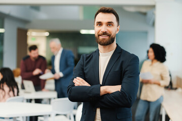 Portrait of attractive smiling bearded man, employee with crossed arms working in modern office