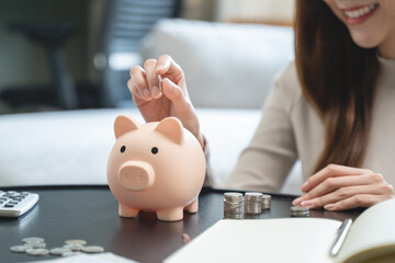 Young woman saving money monthly expenses putting coin in to piggy bank on the table.