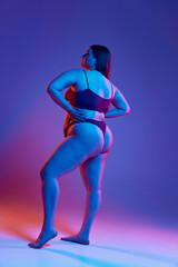 Full-length back view image of young girl posing in underwear, with plus size body shape against purple studio background in neon light. Concept of natural beauty, body positivity, care, acceptance