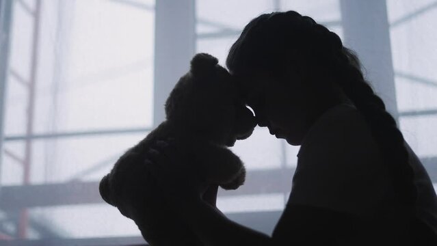 depressed girl child silhouette the window with her favorite toy teddy bear upset. problem of domestic violence in the family lifestyle concept. depressed girl is sad and bored at window with bear