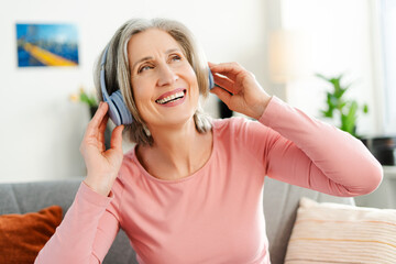 Naklejka premium Overjoyed senior woman listening music in wireless headphones, having fun sitting on sofa