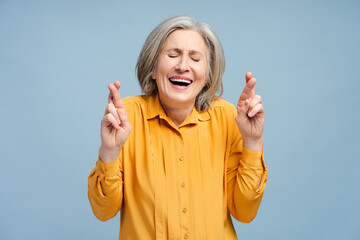 Smiling senior woman wearing casual shirt with crossed fingers standing isolated