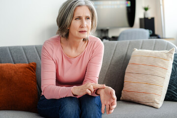 Sad senior woman touching hand, measuring blood pressure, sitting on comfortable sofa