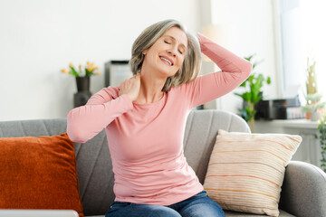 Smiling active senior woman stretching head, doing exercises sitting on comfortable sofa