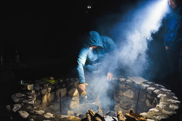 A man with a mysterious air stands before a crackling fire pit, smoke billowing around him in...