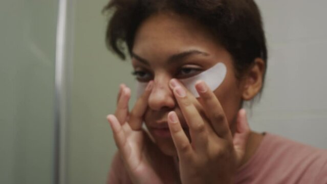 Close up view of drowsy girl with swollen face applying eye patches and looking in bathroom mirror while feeling unwell in morning