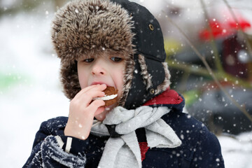 Portrait of a little boy in a fur hat eating cookies on a winter day.