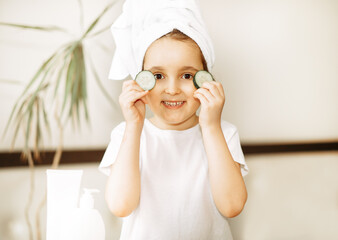 Funny little girl with towel on her head, holding two slices of fresh cucumber and looking at camera