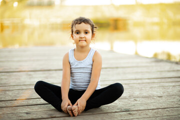 Little girl sitting in nature near by river lake and practicing yoga or resting after sports exercises. Healthy lifestyle.