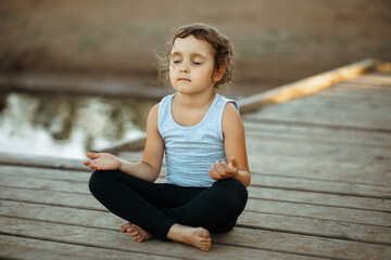 Llittle girl sitting in nature near by river or lake and practicing yoga. Healthy lifestyle.