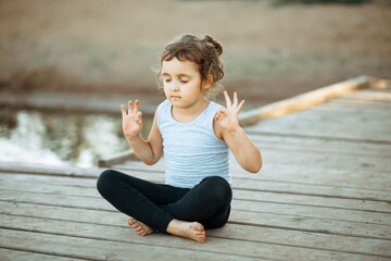 Llittle girl sitting in nature near by river or lake and practicing yoga. Healthy lifestyle.