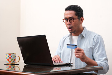 A man looking to his laptop showing shocked expression while holding credit card
