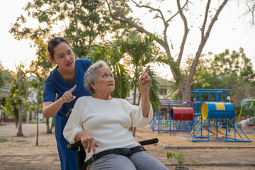 Young nurse and elderly asian senior woman on wheelchair, Asian careful caregiver and patient,  Nurse gives a pep talk to an elderly female patient in the garden in the evening. Health insurance.