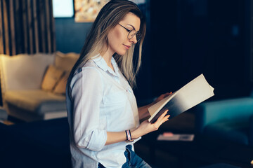 Gorgeous contemplative intelligent woman in glasses for eyes protection reading articles in popular magazine sitting on sofa.Pretty pensive hipster girl enjoying recreation and viewing photos indoors
