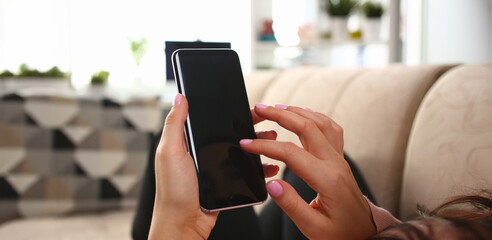 Young beautiful woman holds smartphone in hand using at home costs about sofa. Verifies internal...