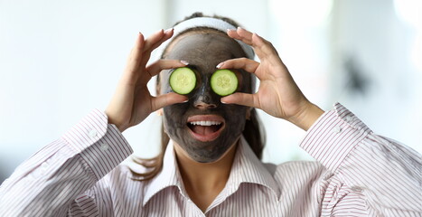 Woman makes gray clay facial mask at home. Put cut cucumber on her eyes like glasses. Spa home...
