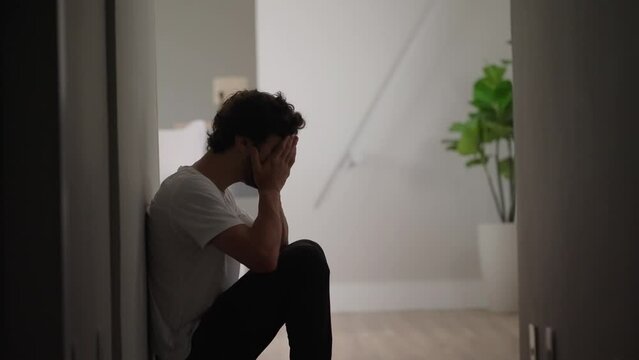Sad young Man Sliding Down the Wall of Her Apartment, Covering Face with Hands. Atmosphere of Depression, Sickness, Tragedy, Death in the Family. Dramatic Bad News Cinematic Slow Motion Moment

